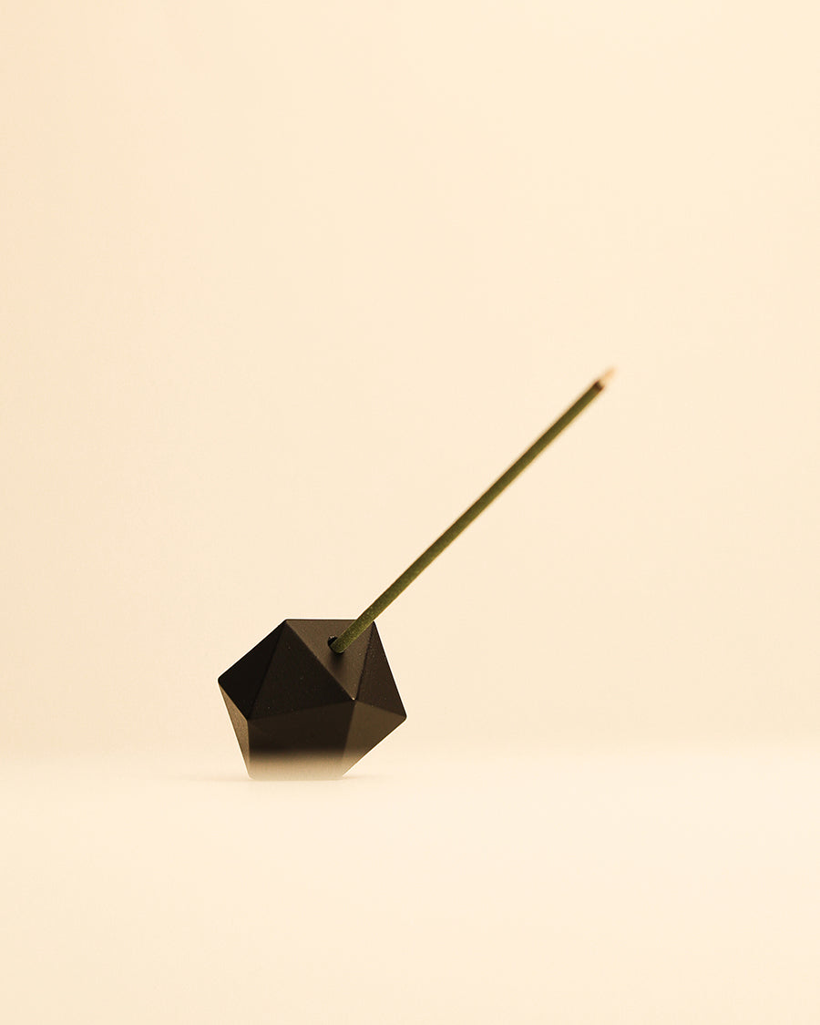 Minimalist matte black incense holder resting on a tabletop. Black geometric incense holder with a lit green incense stick on a beige background